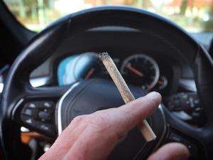 Ein Mann sitzt mit einem Joint zwischen den Fingern am Steuer eines Autos. 