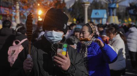 24.01.2023, China, Peking: Ein Mann mit einer Gesichtsmaske hält gegrillten Tintenfisch, während die Besucher eines Tempelmarktes während des einwöchigen Neujahrsfestes vorbeischauen. Foto: Andy Wong/AP/dpa +++ dpa-Bildfunk +++