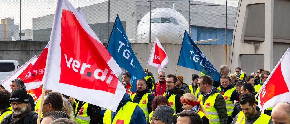 Ein Protestzug von Streikenden zieht mit Bannern und Verdi-Fahnen vom Lufthansa Aviation Center zum Terminal 1. Im Hintergrund ist ein Flugzeug der Lufthansa zu sehen.
