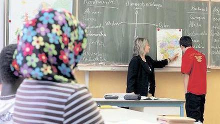 Eine Lehrerin erklärt einem Schüler an der Tafel eine Landkarte. Im Vordergrund sitzen ein weiterer Schüler mit dunkler Hautfarbe und eine Schülerin mit Kopftuch.