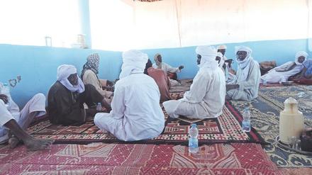 Männer in meist weißen Gewändern und mit turbanartigen Kopfbedeckungen sitzen in einem großen Raum auf Teppichen und reden miteinander.