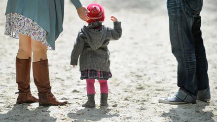 Ein Kleinkind steht mit zwei Erwachsenen auf einem Spielplatz im Sand.