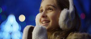 Close up of the happy cute girl looking charmed at something and clapping in white winter gloves on the blurred Christmas lights in the night. Portrait shot. Outside