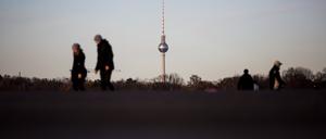 Spaziergänger gehen auf dem Tempelhofer Feld vor dem Berliner Fernsehturm, wobei sich die Szenerie in einer Pfütze spiegelt.
