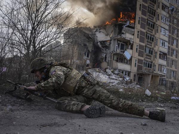 ARCHIV - 17.03.2023, Ukraine, Awdijiwka: Dieses von der Nachrichtenagentur AP am 21.03.2023 zur Verfügung gestellte Foto zeigt einen ukrainischen Polizisten, der vor einem brennenden Gebäude in Deckung geht. 