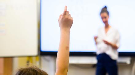 Ein Schüler meldet sich per Handzeichen während eine Lehrerin vor einer digitalen Schultafel steht. 