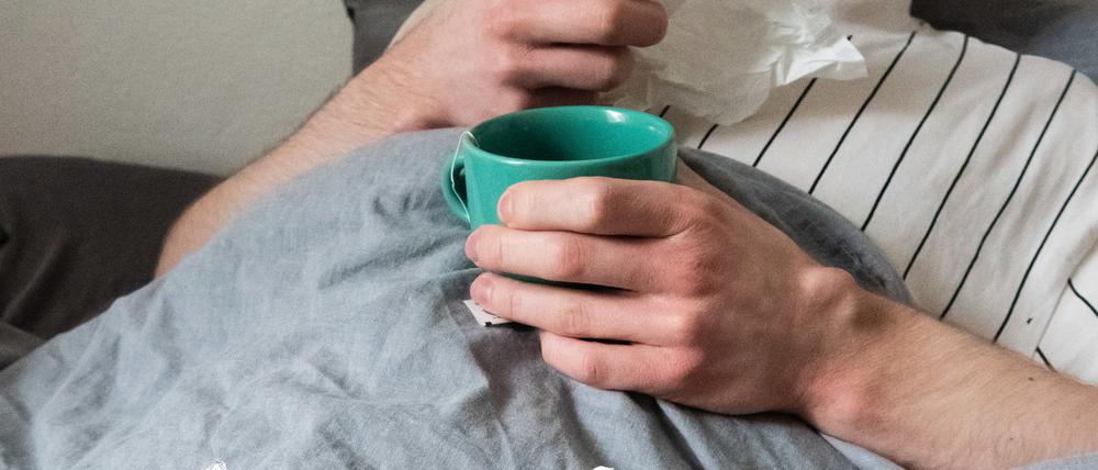 Ein Mann liegt mit Taschentüchern, Teetasse, Nasenspray und Tabletten im Bett (gestellte Szene). (Symbolbild)