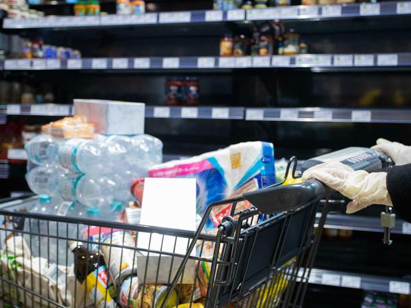 Ein Kunde des Supermarkts schiebt seinen vollen Einkaufswagen mit Hygienehandschuhen an einem leeren Regal vorbei.