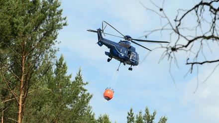 Ein Hubschrauber mit angehängtem Wassertank fliegt über die Bäume eines Waldstücks. 