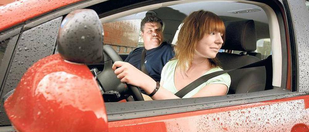 Eine junge Frau sitzt in einem roten Auto und schaut über ihre Schulter, neben ihr ein älterer Mann. 