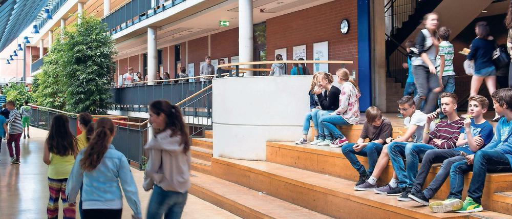 Das frisch sanierte Foyer einer Ganztagsschule, in dem Schüler auf Holzstufen sitzen und Schülerinnen an begrünten Flächen vorbeigehen.