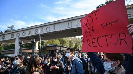 Studierende mit Mundschutz stehen vor einem Universitätsgelände, einer hält ein Plakat mit der Aufschrift Not my rector hoch.